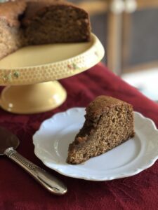 A slice of jam cake on a plate, with the rest of the cake in the background