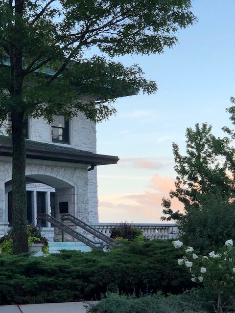 Pink clouds and a blue sky glow in the distance beyond Piper Hall's main stairs, with green foliage in bloom in the foreground.