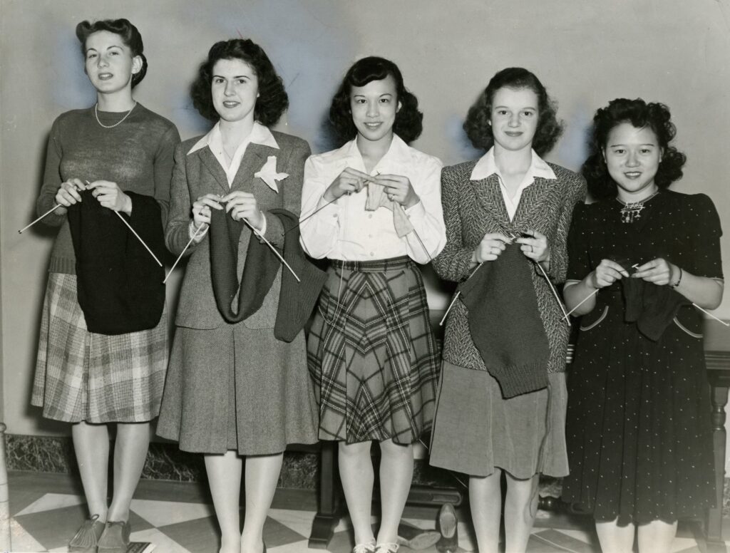 Five Mundelein College students dressed in 1940s fashions stand in a line and hold their knitting projects.