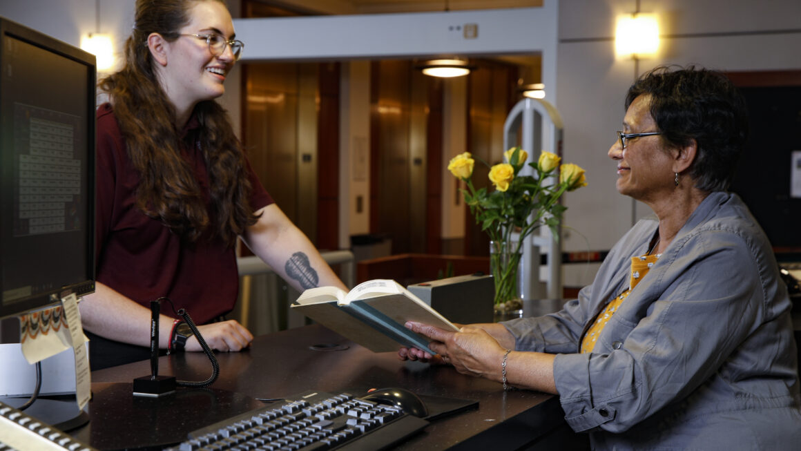 person standing in front of the Circulation Desk while the faculty librarian helps them with checking out a book