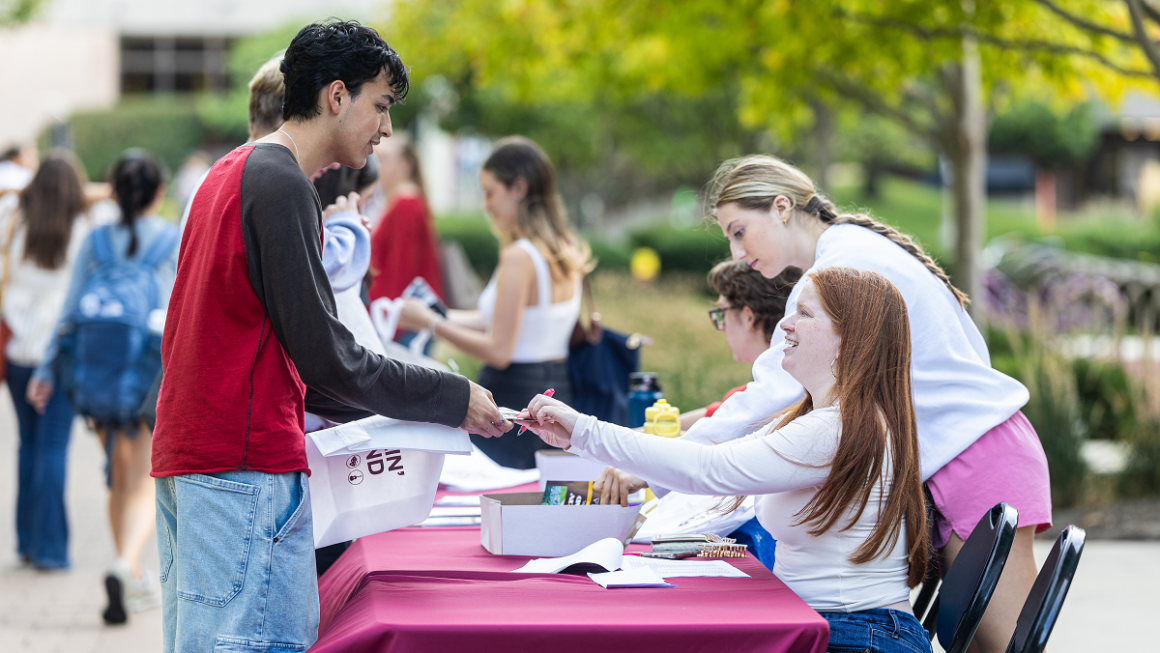 student standing in front of a table with maroon cloth and person sitting behind the table is handing materials