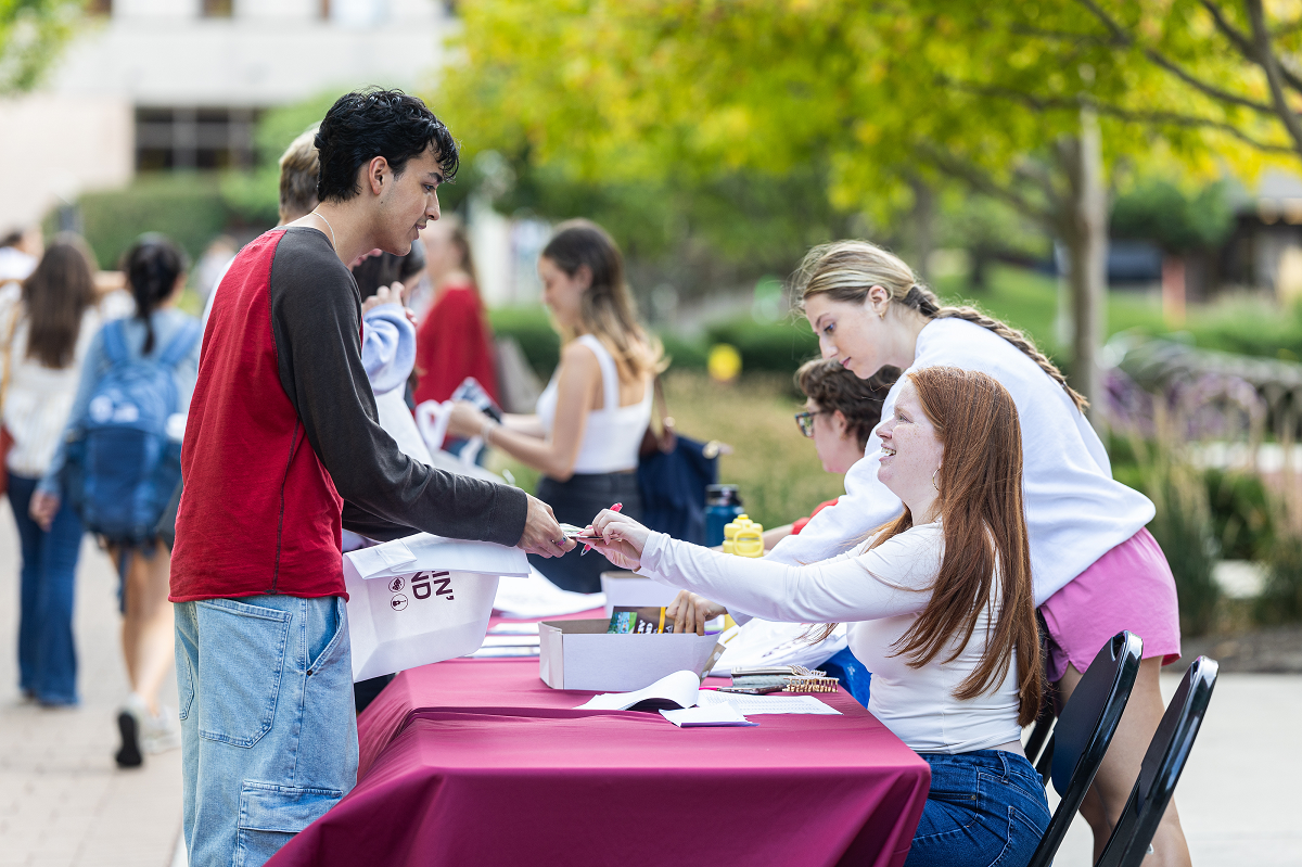 student standing in front of a table with maroon cloth and person sitting behind the table is handing materials